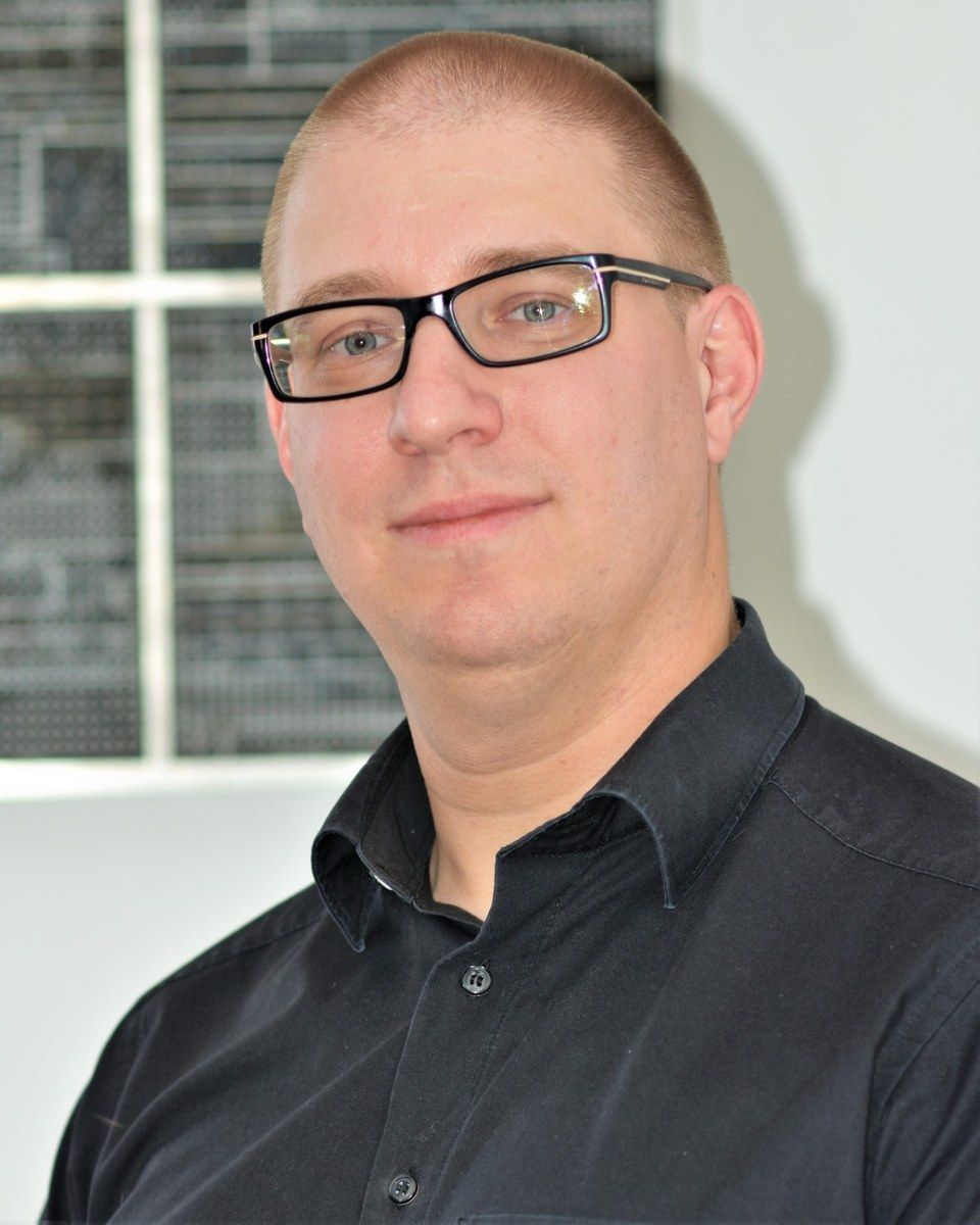 Porträt von Jens Paul: Brustbild mit dunkler Brille und schwarzem Hemd vor heller Wand